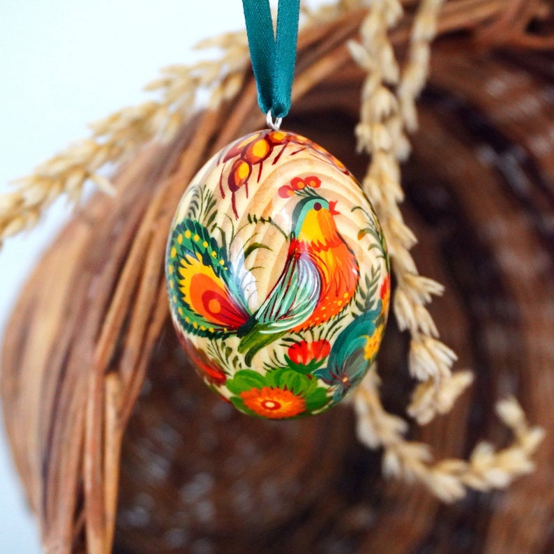 May include: A hand-painted wooden Easter egg with a colorful rooster design. The egg is decorated with flowers and leaves in shades of red, green, yellow, and orange.