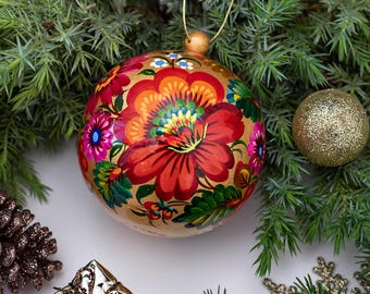 May include: A spherical Christmas bauble featuring a hand-painted floral design with large red, orange, and yellow petals, smaller pink and blue flowers, and green leaves on a gold background, suspended by a thin gold string.