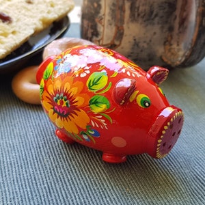 Peut inclure: Une tirelire en céramique rouge avec un motif floral peint dessus. Les fleurs sont jaunes, orange et bleues. La tirelire a un petit trou en haut pour les pièces de monnaie.