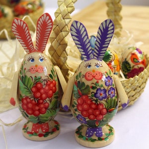 May include: Two wooden Easter eggs decorated with colorful floral and berry patterns. The eggs have bunny ears and are standing upright on a wooden base.