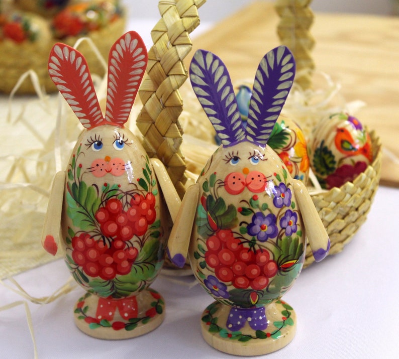 May include: Two wooden Easter eggs decorated with colorful floral and berry patterns. The eggs have bunny ears and are standing upright on a wooden base.