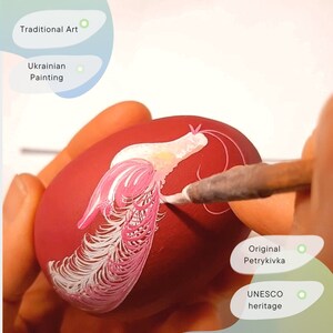 May include: A hand paints a pink and white design on a red egg. The design is a traditional Ukrainian folk art called Petrykivka, which is a UNESCO heritage.