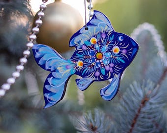May include: A blue fish-shaped Christmas ornament with a floral design in shades of blue, purple, and yellow. The ornament is hanging from a silver string and is displayed on a Christmas tree.