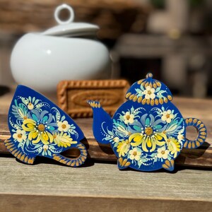 Könnte beinhalten: Zwei handbemalte Holz-Teetassen, eine in Form einer Teekanne und die andere in Form einer Teetasse. Beide sind in einem blauen und weißen Blumenmuster mit goldenen Akzenten bemalt.