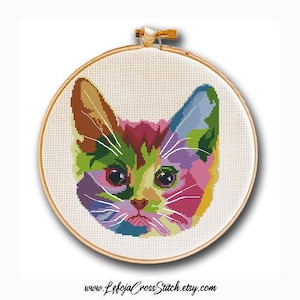 May include: A colorful cross-stitch pattern of a cat's face. The cat's fur is a mix of pink, purple, green, yellow, and blue. The cat's eyes are black with a white highlight. The cat's nose is pink. The cat's whiskers are white.