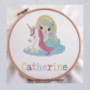 May include: Cross stitch pattern featuring a mermaid with rainbow hair and a unicorn, with the name "Catherine" stitched below. The pattern is displayed in a wooden embroidery hoop.