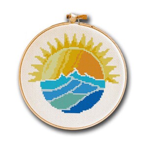 May include: Cross-stitch embroidery in a wooden hoop. The design features a yellow and orange sun setting over blue and teal waves. The embroidery is on a cream-colored fabric.