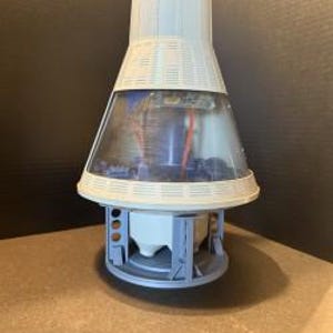 May include: A model of a spacecraft capsule, featuring a white conical top and a transparent, angled midsection. The base is gray with structural supports. Interior details are visible through the clear section, and the model sits on a brown surface.