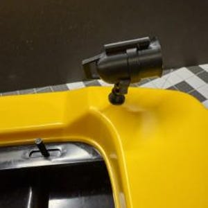 May include: A black spotlight is mounted on a bright yellow surface. The spotlight is cylindrical with a rectangular top and is attached to a swivel base. The yellow surface appears to be part of a larger object, possibly a vehicle or equipment.
