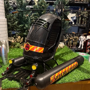 May include: Black toy helicopter with the text "GLIDE CAPTURE COPTER" on the side. The helicopter has a red and yellow striped design and a circular logo with the letter "A". The toy is on a green surface.