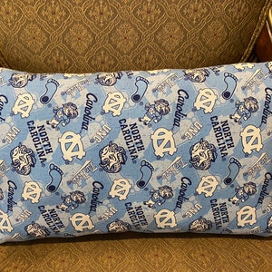 May include: A blue and white decorative pillow with a repeating pattern of the University of North Carolina Tar Heels logo and the words "North Carolina" and "UNC".