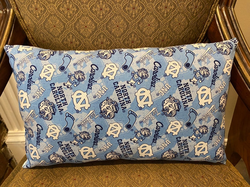 May include: A blue and white decorative pillow with a repeating pattern of the University of North Carolina Tar Heels logo and the words "North Carolina" and "UNC".