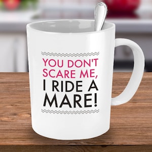 May include: White ceramic coffee mug with black text that reads "You don't scare me, I ride a mare!" in pink lettering.