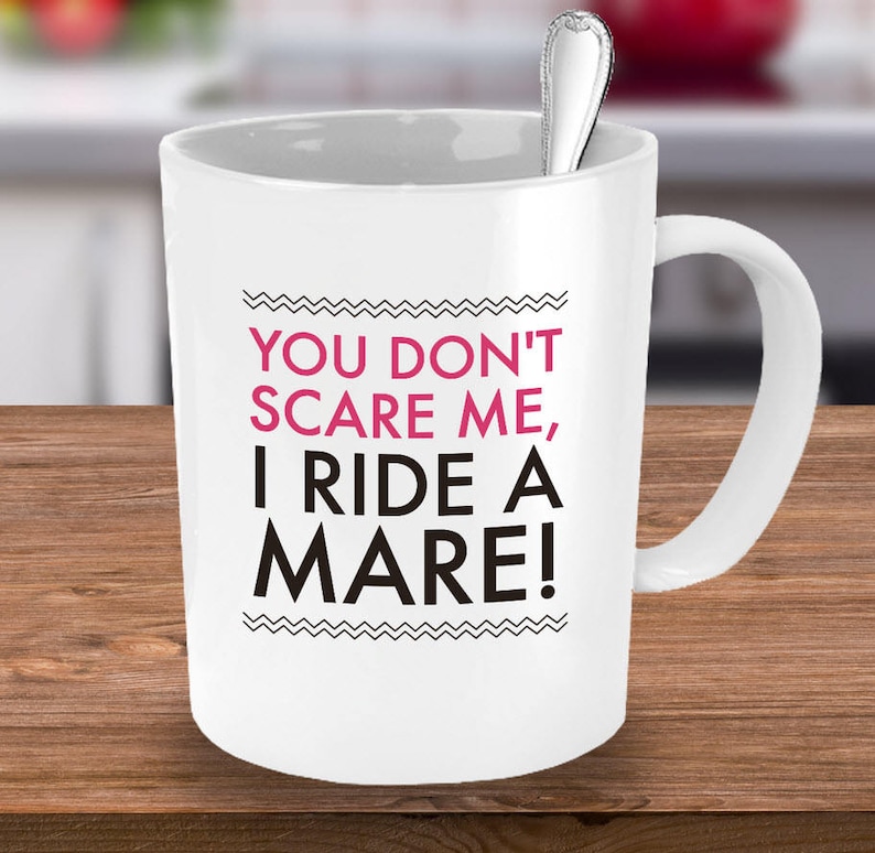 May include: White ceramic coffee mug with black text that reads "You don't scare me, I ride a mare!" in pink lettering.