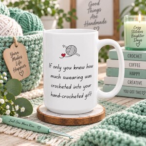 May include: A white ceramic mug featuring a black graphic of a yarn ball with a crochet hook and a small red heart. The text reads: If only you knew how much swearing was crocheted into your hand-crocheted gift. The mug sits on a round coaster.