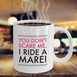 May include: White ceramic coffee mug with black text that reads "You don't scare me, I ride a mare!" The text is outlined with a black wavy line.