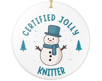 May include: White ceramic Christmas ornament with a snowman design. The snowman wears a blue top hat, scarf, and has black button eyes and a coal mouth. Text reads "CERTIFIED JOLLY KNITTER" in blue.