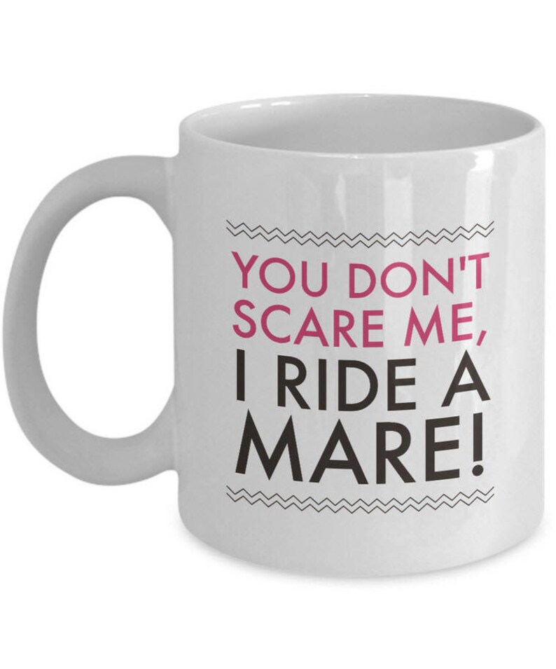 May include: White ceramic coffee mug with a black and white zig zag design around the text "You don't scare me, I ride a mare!" in black with "mare" in pink.