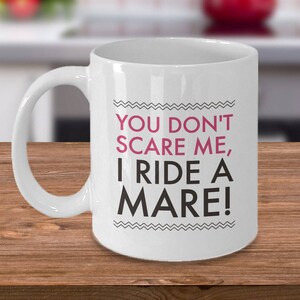 May include: White ceramic coffee mug with a black and white graphic design. The text on the mug reads "You don't scare me, I ride a mare!"