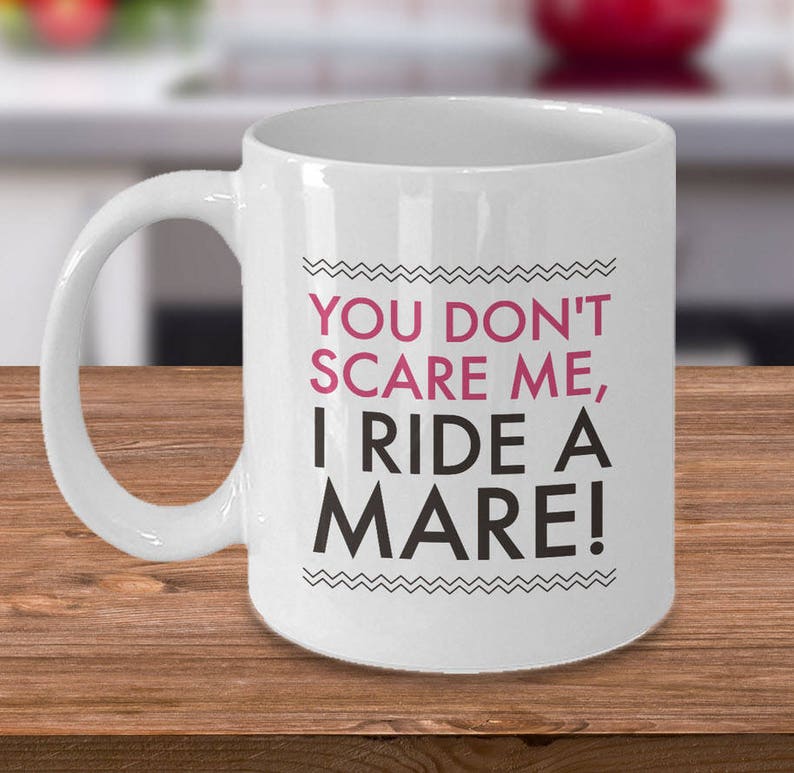 May include: White ceramic coffee mug with a black and white graphic design. The text on the mug reads "You don't scare me, I ride a mare!"