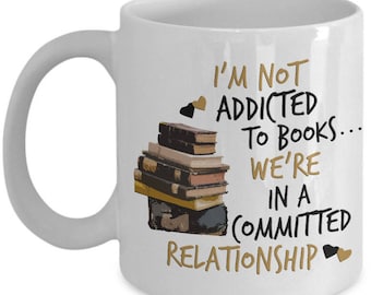 Puede incluir: Taza de cerámica blanca con asa curva. La taza presenta una pila de libros y el texto "I'M NOT ADDICTED TO BOOKS... WE'RE IN A COMMITTED RELATIONSHIP" en letras negras y doradas. También se incluyen dos pequeños gráficos de corazón.