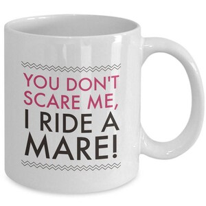 May include: White ceramic coffee mug with black text that reads "You don't scare me, I ride a mare!" The text "mare" is in pink.