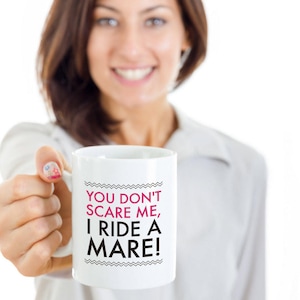 May include: White ceramic coffee mug with black text that reads "You don't scare me, I ride a mare!" in pink lettering.
