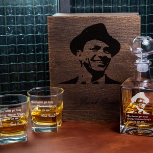 Puede incluir: Una caja de madera con una imagen en blanco y negro de Frank Sinatra en la parte delantera. La caja contiene dos vasos de vidrio transparente y una jarra de vidrio transparente con un tapón. Los vasos y la jarra están grabados con la cita "You better get busy living, because dying's a pain in the ass." - Frank Sinatra.