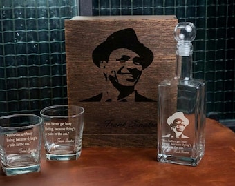 May include: A clear glass decanter with a spherical stopper and two matching square whiskey glasses, all etched with a portrait of Frank Sinatra and the quote, "You better get busy living, because dying's a pain in the ass." A wooden box features the same portrait.