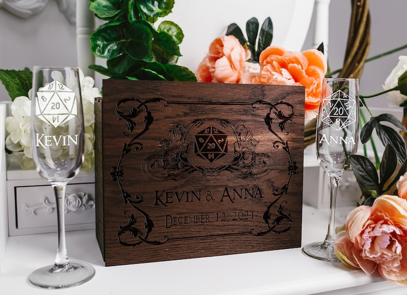 May include: A wooden box with a 20-sided die design and the text "Kevin & Anna December 12, 2021". Two champagne flutes with 20-sided die designs are on either side of the box. The flutes are engraved with the names "Kevin" and "Anna".