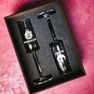 May include: Two champagne flutes in a black box with foam padding. The flutes are engraved with the names "Markus" and "Jenny" and a star wars inspired design.