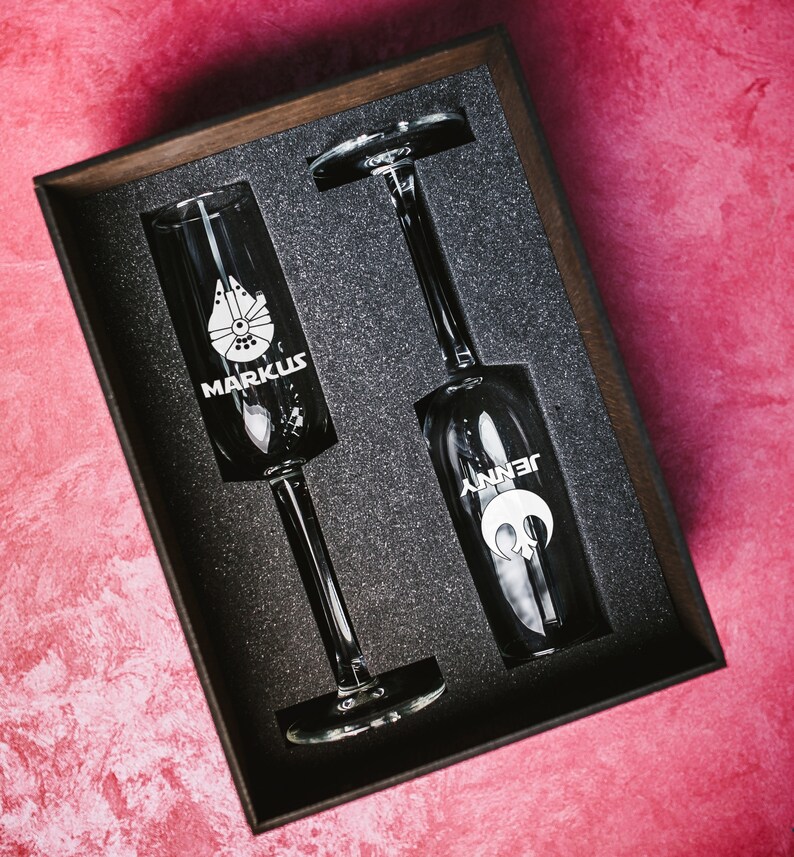 May include: Two champagne flutes in a black box with foam padding. The flutes are engraved with the names "Markus" and "Jenny" and a star wars inspired design.