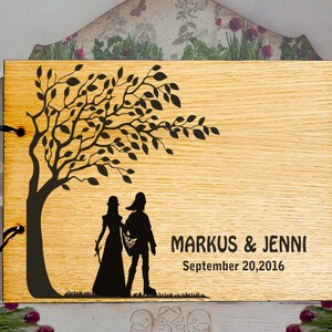 May include: A wooden photo album with a laser-cut design of a couple standing under a tree. The album has a black ribbon closure and the text "MARKUS & JENNI September 20, 2016" is engraved on the front.