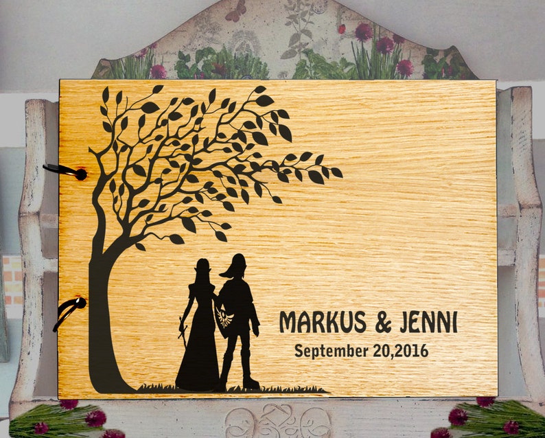 May include: A wooden photo album with a laser-cut design of a couple standing under a tree. The album has a black ribbon closure and the text "MARKUS & JENNI September 20, 2016" is engraved on the front.