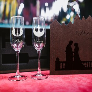 May include: Two champagne flutes with engraved Star Wars symbols and the names "Paolo" and "Sara". A wooden box with a silhouette of a couple and the text "Paolo & Sara 17 September 2022" is in the background.