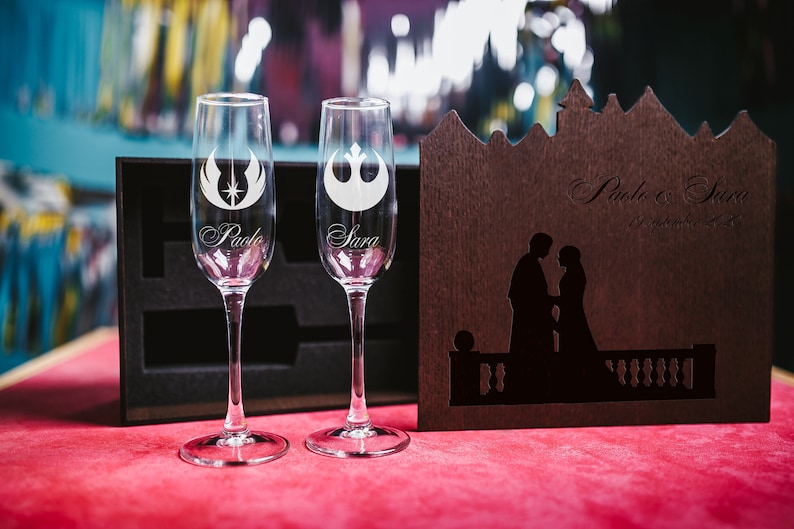 May include: Two champagne flutes with engraved Star Wars symbols and the names "Paolo" and "Sara". A wooden box with a silhouette of a couple and the text "Paolo & Sara 17 September 2022" is in the background.