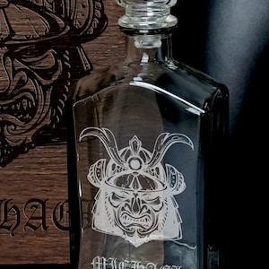 May include: A clear glass decanter with a square stopper, featuring an etched samurai helmet design and the name MICHAEL engraved on the front. The decanter is positioned in front of a dark wooden box that also displays a matching samurai helmet engraving.