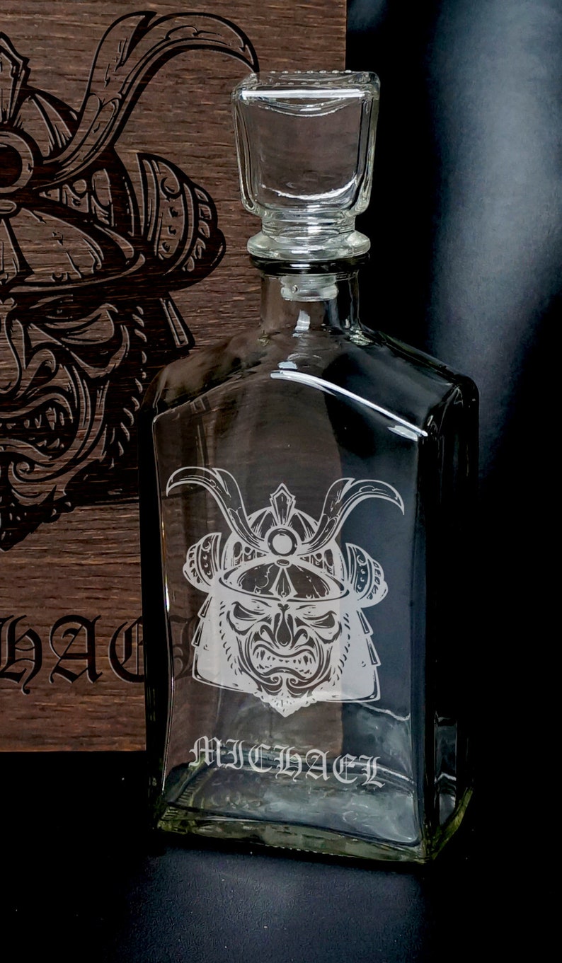 May include: A clear glass decanter with a square stopper, featuring an etched samurai helmet design and the name MICHAEL engraved on the front. The decanter is positioned in front of a dark wooden box that also displays a matching samurai helmet engraving.