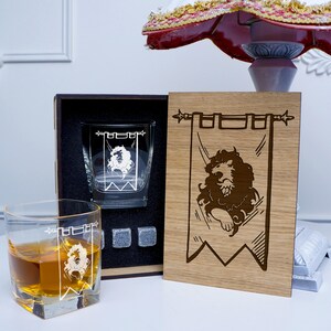 May include: A wooden box set with two engraved whiskey glasses and a wooden plaque with a lion emblem. The glasses are clear and have a lion design. The plaque is brown and has the words "House Lannister" engraved on it.