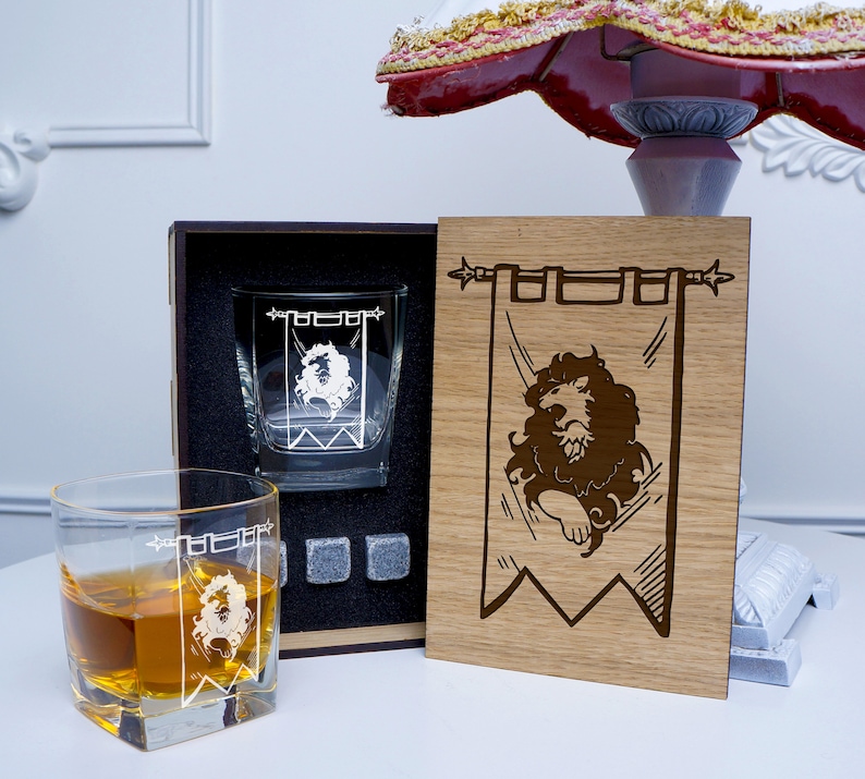 May include: A wooden box set with two engraved whiskey glasses and a wooden plaque with a lion emblem. The glasses are clear and have a lion design. The plaque is brown and has the words "House Lannister" engraved on it.