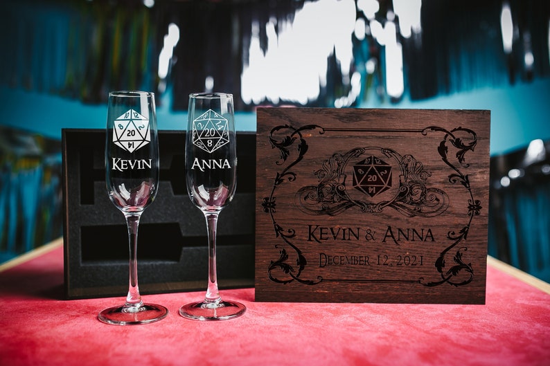 May include: Two engraved champagne flutes with a 20-sided die design and the names Kevin and Anna. The flutes are in a wooden box with a similar design and the text "Kevin & Anna December 12, 2021".