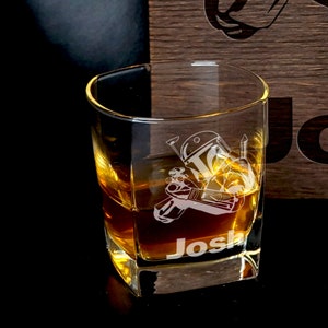 Puede incluir: Un vaso cuadrado de vidrio transparente con un dise&ntilde;o de Boba Fett y el nombre "Josh" grabado en la parte inferior. El vaso est&aacute; lleno de l&iacute;quido &aacute;mbar.