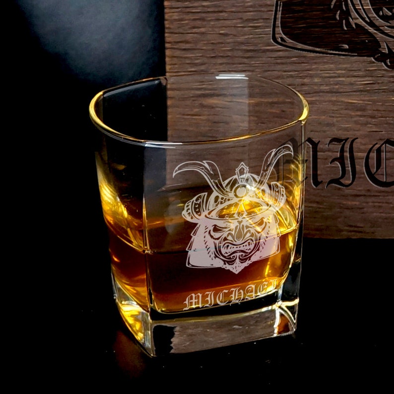 May include: A clear glass tumbler filled with amber liquid, featuring an etched samurai helmet design and the name MICHAEL engraved below it. The glass has a thick base and sits in front of a dark wooden box with partial text visible.