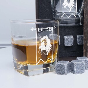 May include: A set of four clear glass whiskey glasses with a white graphic of a lion in a banner. The glasses are filled with amber liquid and are sitting on a black surface with gray stone coasters.