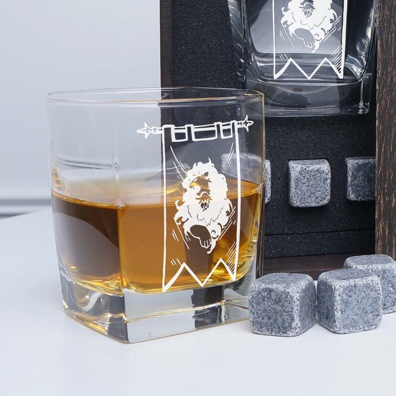 May include: A set of four clear glass whiskey glasses with a white graphic of a lion in a banner. The glasses are filled with amber liquid and are sitting on a black surface with gray stone coasters.