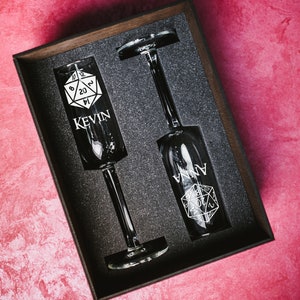 May include: Two champagne flutes with a black and white dice design and the names "Kevin" and "Anna" engraved on them. The flutes are in a black box with a foam interior.