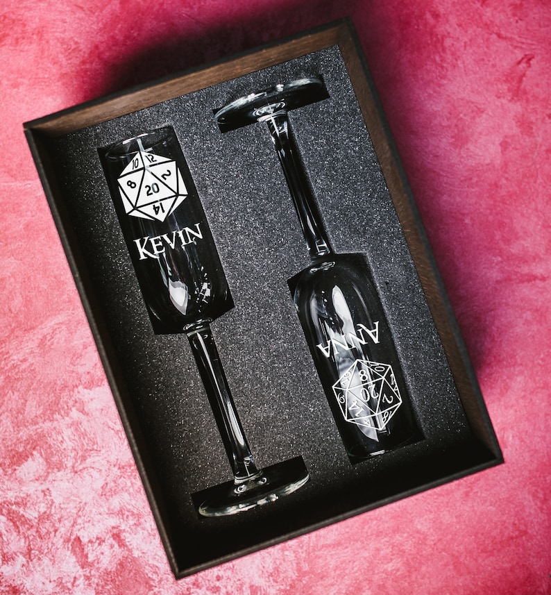 May include: Two champagne flutes with a black and white dice design and the names "Kevin" and "Anna" engraved on them. The flutes are in a black box with a foam interior.