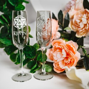 May include: Two champagne flutes with a 20-sided die design and the names "Kevin" and "Anna" etched on them. The flutes are on a white surface with pink flowers and green leaves.