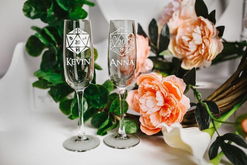 May include: Two champagne flutes with a 20-sided die design and the names "Kevin" and "Anna" etched on them. The flutes are on a white surface with pink flowers and green leaves.