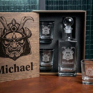May include: A wooden gift box containing a clear glass decanter and three matching square glasses. The box lid and each glass feature an engraved samurai helmet graphic and the name Michael. The decanter has a round glass stopper.